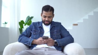 Neşeli Afrikalı Amerikalı adam evde akıllı telefon kullanıyor ve gülüyor. Sandalyeye oturmuş telefona bakıyor ve internette yazı yazıyor.
