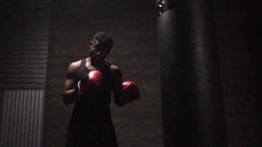 Afrika kökenli Amerikalı genç boksör. Koyu renk spor salonunda antrenman yapıyor. Eldivenlerle kum torbasına vuruyor.