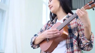 Genç Asyalı kadın evde durup ukulele çalıyor ve hayal kuruyor, bayan müzisyen akustik gitar çalıyor ve hayal ediyor.