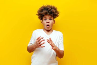 Korkmuş kıvırcık Afrikalı Amerikalı çocuk sarı izole edilmiş arka planda korku ve şok belirtisi gösteriyor. Beyaz tişörtlü genç şaşırmış bir şekilde fotokopi odasına bakıyor.