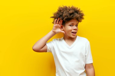 Sarı izole edilmiş arka planda kulak misafiri olan, beyaz tişörtlü, gözetleyen ve dinleyen, mütevazı genç, meraklı kıvırcık Afrikalı Amerikalı çocuk.