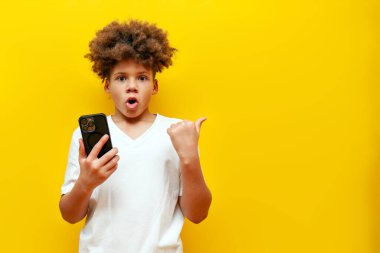 Şok olmuş Afrikalı Amerikalı çocuk akıllı telefon kullanıyor ve sarı izole edilmiş arka plandaki kopya alanını işaret ediyor, şaşırmış okul çocuğu cep telefonu tutuyor ve şaşkınlıkla boş alanı işaret ediyor.