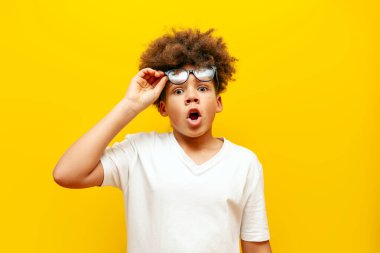 Şaşırmış kıvırcık saçlı gözlüklü Amerikalı çocuk sarı izole edilmiş arka planda şoka girmiş beyaz tişörtlü genç şaşkınlıkla fotokopi odasına bakıyor.