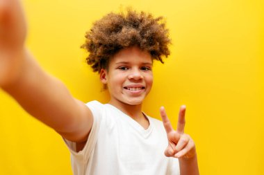 Neşeli Afrikalı Amerikalı çocuk selfie çekiyor ve sarı izole edilmiş arka planda barış işareti gösteriyor. Kıvırcık okul çocuğu akıllı telefondan fotoğraf çekiyor ve iki parmağını gösteriyor.