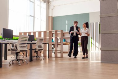 Afrika kökenli Amerikalı kadın ve Avrupalı takım elbiseli genç adam ofiste yürüyor ve raporları ve istatistikleri tartışıyor, ofis çalışanları kağıtlarla konuşuyor ve çalışıyor.