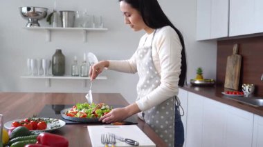 Önlüklü genç Asyalı esmer kadın salata hazırlıyor ve mutfakta tabakta malzemeleri karıştırıyor. Koreli ev hanımı evde sağlıklı vejetaryen yemekleri hazırlıyor.