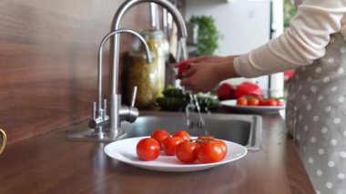 Önlüklü Asyalı genç bir kadın mutfak lavabosunda kırmızı domates yıkıyor, ev hanımı evde salata hazırlıyor ve sebzeleri suyla soyuyor.