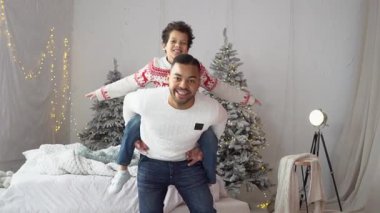 Afrika kökenli Amerikalı bir baba Noel kıyafetleri içinde oğluyla eğleniyor, ebeveynlerinin sırtına oturmuş ve uçuyormuş gibi yapıyor, Noel 'i evde kutluyor ve dans ediyor.