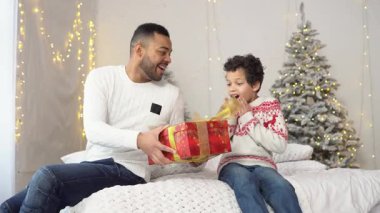 Afrikalı Amerikalı baba, yeni yıl için oğluna hediye veriyor, çocuk Noel için hediye kutusunu açıyor ve mutlu ve şaşırıyor, aile yeni yıl kıyafetleriyle Noel 'i kutluyor.