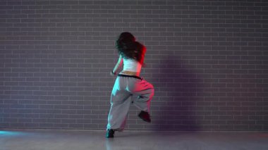 Genç kız koreograf dans ediyor, neon ışıkta hip hop yapıyor, kamerada şarkı söyleyen kadın karanlık stüdyoda, arka planda dans ediyor.