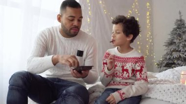 Yılbaşı arifesinde Noel ağacının yanında tatlı çikolatalı kurabiyeler yiyen Afrikalı Amerikalı baba ve oğul Noel 'i evde birlikte kutluyor ve tatlı yiyorlar.