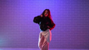 Genç kız koreograf dans ediyor, neon ışıkta hip hop yapıyor, kadın kamera önünde şarkı söylüyor ve karanlık stüdyoda dans çalışıyor.