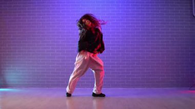 Genç kız koreograf dans ediyor, neon ışıkta hip hop yapıyor, kadın kamera önünde şarkı söylüyor ve karanlık stüdyoda dans çalışıyor.