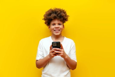 Beyaz tişörtlü neşeli Afro-Amerikalı çocuk. Akıllı telefon kullanıyor ve sarı izole edilmiş arka planda gülümsüyor. Kıvırcık okul çocuğu internette mesaj yazıyor ve cep telefonuna bakıyor.