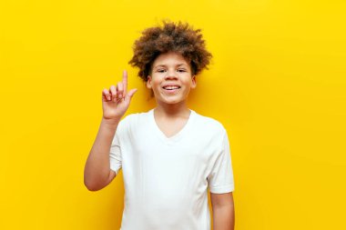 Neşeli kıvırcık Afrikalı Amerikalı çocuk, sarı izole edilmiş arka planda parmakla işaret ediyor ve gülümsüyor, beyaz tişörtlü genç yukarıdan fotokopi alanı gösteriyor ve reklamını yapıyor.