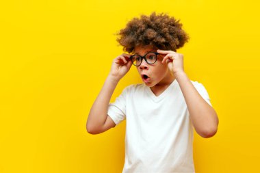 Şaşırmış kıvırcık saçlı, gözlüklü Amerikalı çocuk sarı izole arka planda şoka girmiş, beyaz tişörtlü genç şaşkınlıkla fotokopi odasına bakıyor.