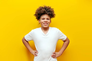 Beyaz tişörtlü neşeli Afro-Amerikan kıvırcık çocuk sarı izole arka planda duruyor, kıvırcık ergen gülümsüyor ve kameraya bakıyor.