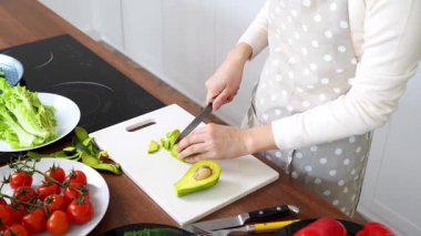 Önlüklü genç Asyalı kadın mutfakta bıçakla avokado kesiyor ve evde yemek pişiriyor. Kız vejetaryen salatasına sebze ve malzeme ekliyor. Yakın plan.