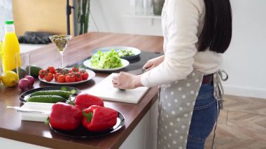 Önlüklü genç Asyalı kadın evde bıçakla salatalık kesiyor ve evde yemek pişiriyor. Kız vejetaryen salatasına sebze ve malzeme ekliyor.