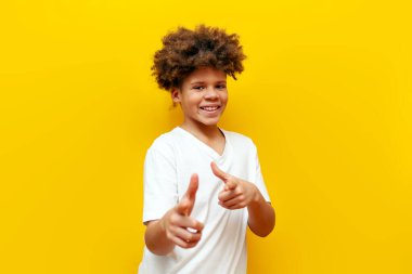 Neşeli Afrikalı Amerikalı çocuk elini öne uzattı ve sarı izole edilmiş arka planda seni seçti. Kıvırcık okul çocuğu kamerayı işaret ediyor ve gülümsüyor.