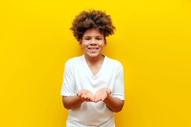 Kıvırcık Afrikalı Amerikalı çocuk elini boş tutuyor ve sarı izole edilmiş arka planı soruyor. Beyaz tişörtlü genç, avuç içlerindeki fotokopi alanını gösteriyor ve alıyor.