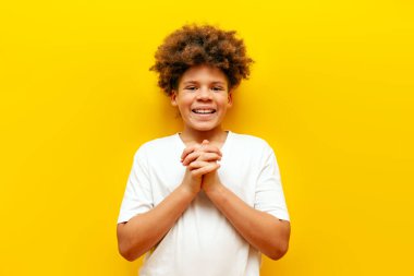 Beyaz tişörtlü Afro-Amerikan kıvırcık çocuk gülümsüyor ve sarı izole arka planda kameraya bakıyor.