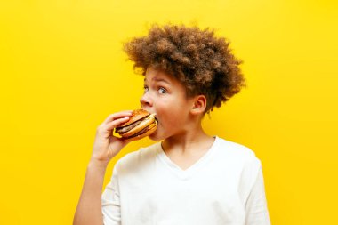Neşeli kıvırcık Afrikalı Amerikalı çocuk sarı izole edilmiş arka planda hamburger yiyor, aç ergen elinde çizburger tutuyor ve fast food reklamı yapıyor.