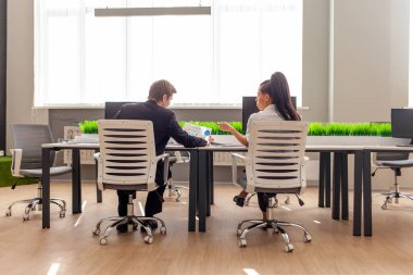 Afrikalı Amerikalı kadın ve Avrupalı takım elbiseli genç adam ofiste oturup rapor ve istatistikleri tartışıyor, ofis çalışanları kağıtlarla konuşuyor ve çalışıyor.