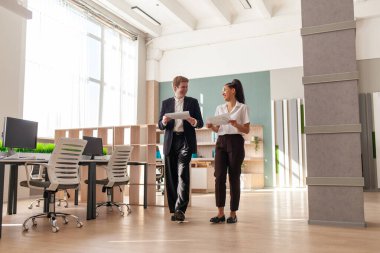 Afrika kökenli Amerikalı kadın ve Avrupalı takım elbiseli genç adam ofiste yürüyor ve raporları ve istatistikleri tartışıyor, ofis çalışanları kağıtlarla konuşuyor ve çalışıyor.