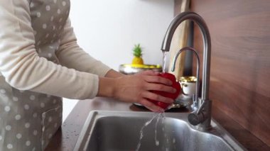 Önlüklü Asyalı genç bir kadın mutfak lavabosunda kırmızı dolma biber yıkıyor, ev hanımı evde salata hazırlıyor ve sebzeleri suyla soyuyor.