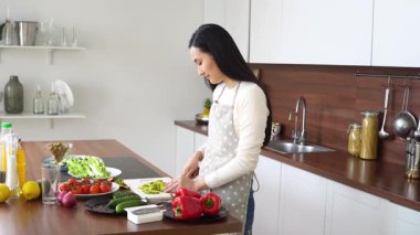 Önlüklü genç Asyalı kadın mutfakta bıçakla avokado kesiyor ve evde yemek pişiriyor. Kız vejetaryen salatasına sebze ve malzeme ekliyor.