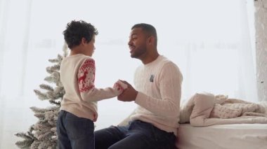 Noel kıyafetleri içinde Afrika kökenli Amerikalı baba ve oğul evde oturuyor ve yılbaşından önce konuşuyor, Noel 'de çocuğa sarılıp gülümsüyor.