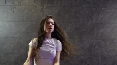 Dans salonundaki gri duvara karşı enerjik bir şekilde dans eden genç ve güzel bir kadın. Şık, modaya uygun bir kız. Hip hop yapıyor ve stüdyoda müzik yapıyor.
