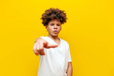 Heyecanlı Afrikalı Amerikalı çocuk elini öne uzattı ve sarı izole edilmiş arka planda suçlamada bulundu. Kıvırcık okul çocuğu kameraya bakıp şaşırdı.