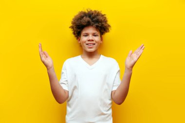Kıvırcık Afrikalı Amerikalı çocuk, sarı izole edilmiş arka planda elleri boş duruyor. Beyaz tişörtlü genç, avuç içlerindeki fotokopi alanını gösteriyor ve alıyor.