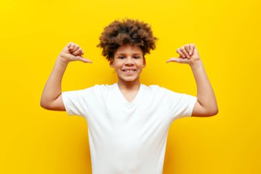 Neşeli Afrikalı Amerikalı çocuk kendini işaret ediyor ve sarı izole edilmiş arka planda gülümsüyor. Kıvırcık okul çocuğu başarıya seviniyor ve zaferi el kaldırarak kutluyor.