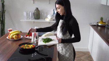 Önlüklü genç bir kadın mutfakta bitkilerle omlet serpiyor. Kızarmış yumurta hazırlıyor. Maydanoz ve soğan ekliyor.