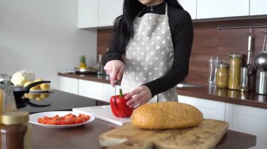 Önlüklü genç bir kadın mutfakta kırmızı biber, evde yemek pişiriyor ve sebze doğruyor.