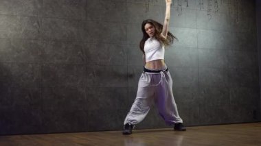 Dans salonundaki gri duvara karşı enerjik bir şekilde dans eden genç ve güzel bir kadın. Şık, modaya uygun bir kız. Hip hop yapıyor ve stüdyoda müzik yapıyor.