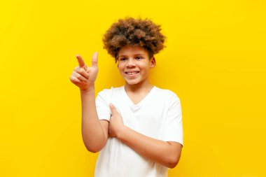 Beyaz tişörtlü Afro-Amerikalı kıvırcık çocuk fotokopi alanını işaret ediyor ve sarı izole arkaplanda boşluğa dokunuyor.