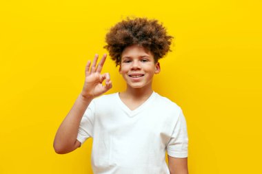 Neşeli kıvırcık Afrikalı Amerikalı çocuk sarı izole edilmiş arka planda jest yapıyor, beyaz tişörtlü genç tavsiye ediyor ve onaylıyor.