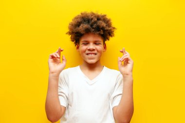 Beyaz tişörtlü Afro-Amerikan kıvırcık saçlı çocuk şans diliyor ve sarı izole arka planda şans diliyor.