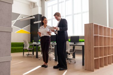 Ofis çalışanları iş kıyafetleri içinde yürüyorlar raporları tartışıyorlar ve ellerinde belgeler tutuyorlar, Afrikalı Amerikalı kadın ve genç Avrupalı erkek toplantıda konuşuyorlar ve planlıyorlar, genel plan