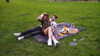 Genç aşık çift parkta battaniyeye uzanır ve akıllı telefondan selfie çeker. Erkek ve kadın, doğadaki temiz havada piknikte dinlenir.