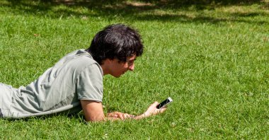 Caucasian young man looking at his smartphone lying on the lawn on a sunny day. Relaxing in the city park
