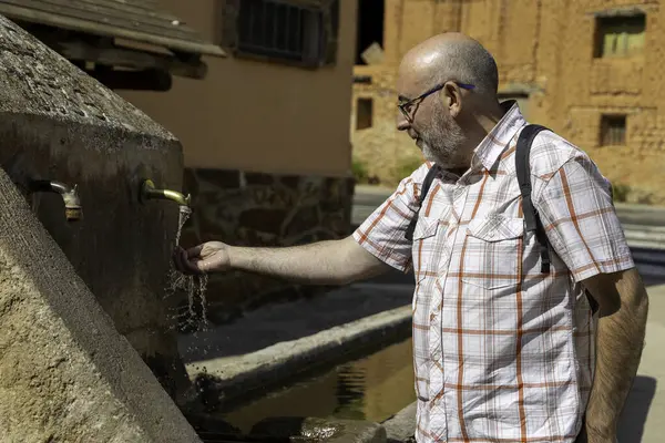 Sakallı yetişkin bir adam köydeki eski bir çeşmeden su topluyor.