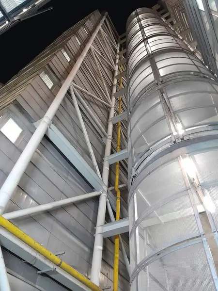 Cylindrical metal structure with horizontal ribs and a wire mesh safety cage, next to a building. There are yellow and white pipes running vertically along the building and structure. The building has a metal exterior with horizontal lines of corruga