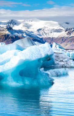 Okyanusta büyük bir buz parçası yüzüyor. Buz arka planda su ve dağlar ile çevrilidir. Sahne huzurlu ve huzurlu, buzlar mavi gökyüzünü yansıtıyor.