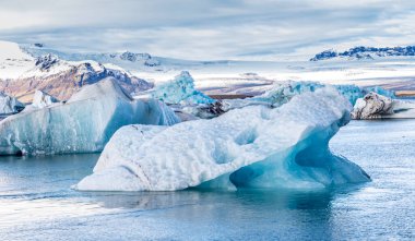Okyanusta yüzen büyük bir buz parçası. Buz daha küçük buz parçalarıyla çevrili. Gökyüzü bulutlu ve su sakin.