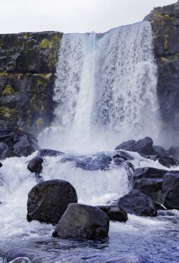 A waterfall with a large rock in front of it. The water is flowing down the rock and the rocks are scattered around the waterfall. The scene is serene and peaceful. The Golden Circle, Iceland. clipart